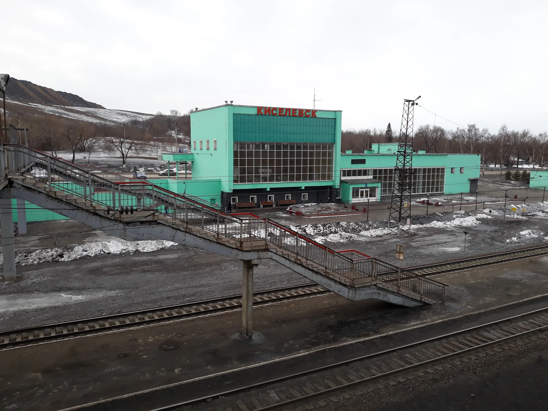 The station building bearing the name Kiselyovsk, railway tracks and a pedestrian bridge; a tall spoil heap is visible in the background on the left.