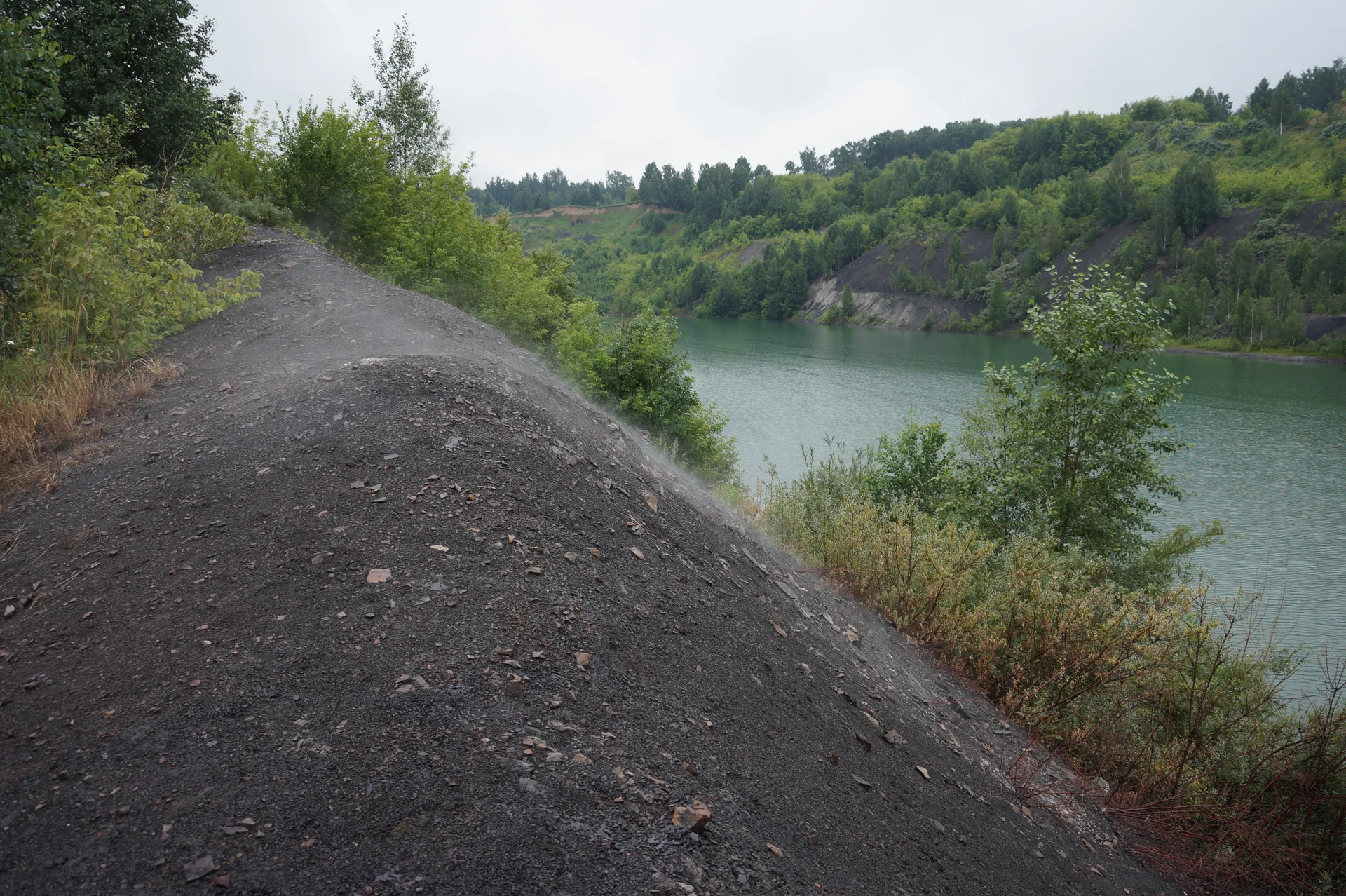 Water between steep slopes; a path follows the bank over dark rock, with trees around it.